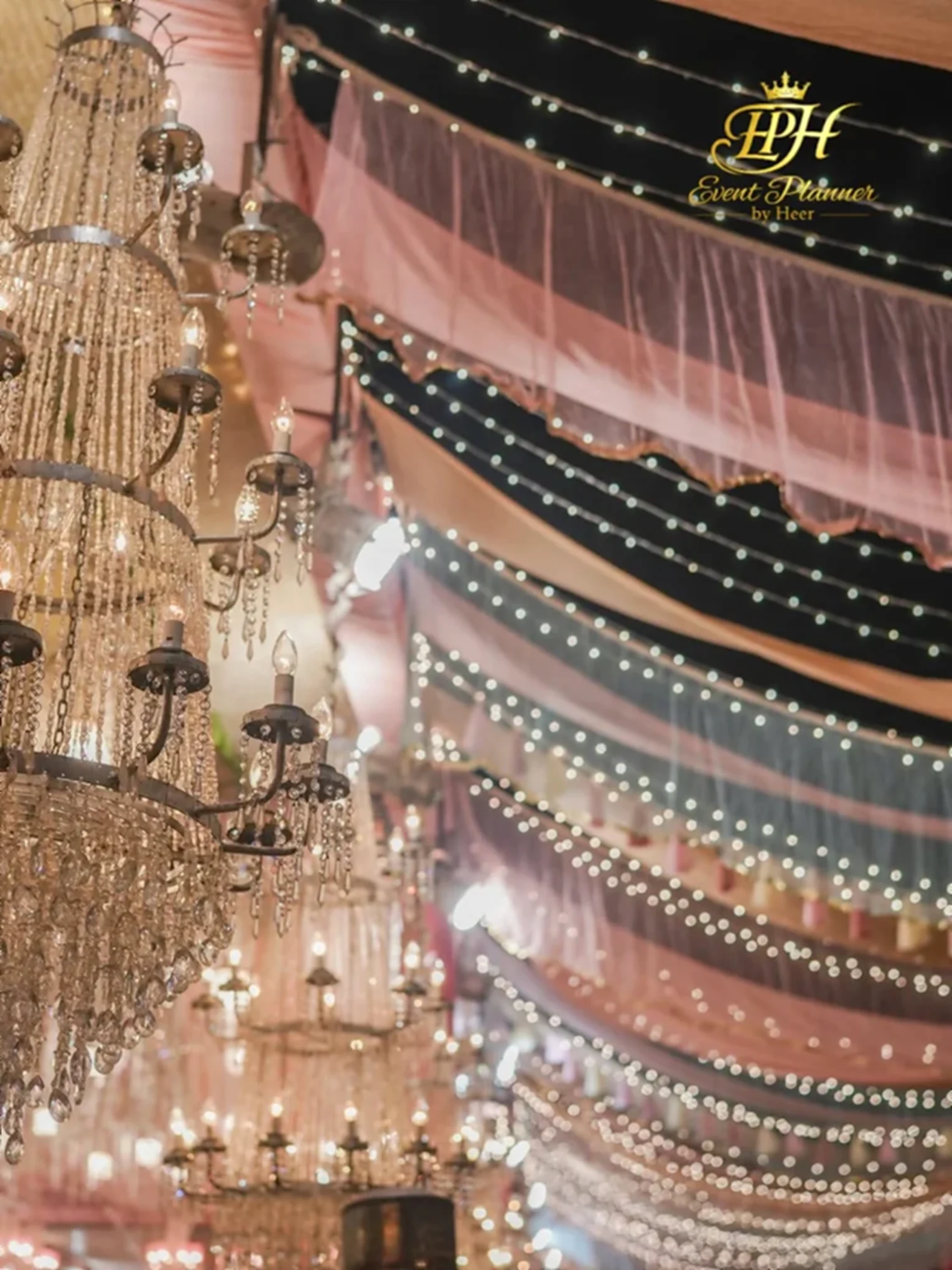 Crystal chandeliers beneath tiered blush and black ceiling draping with strung lights